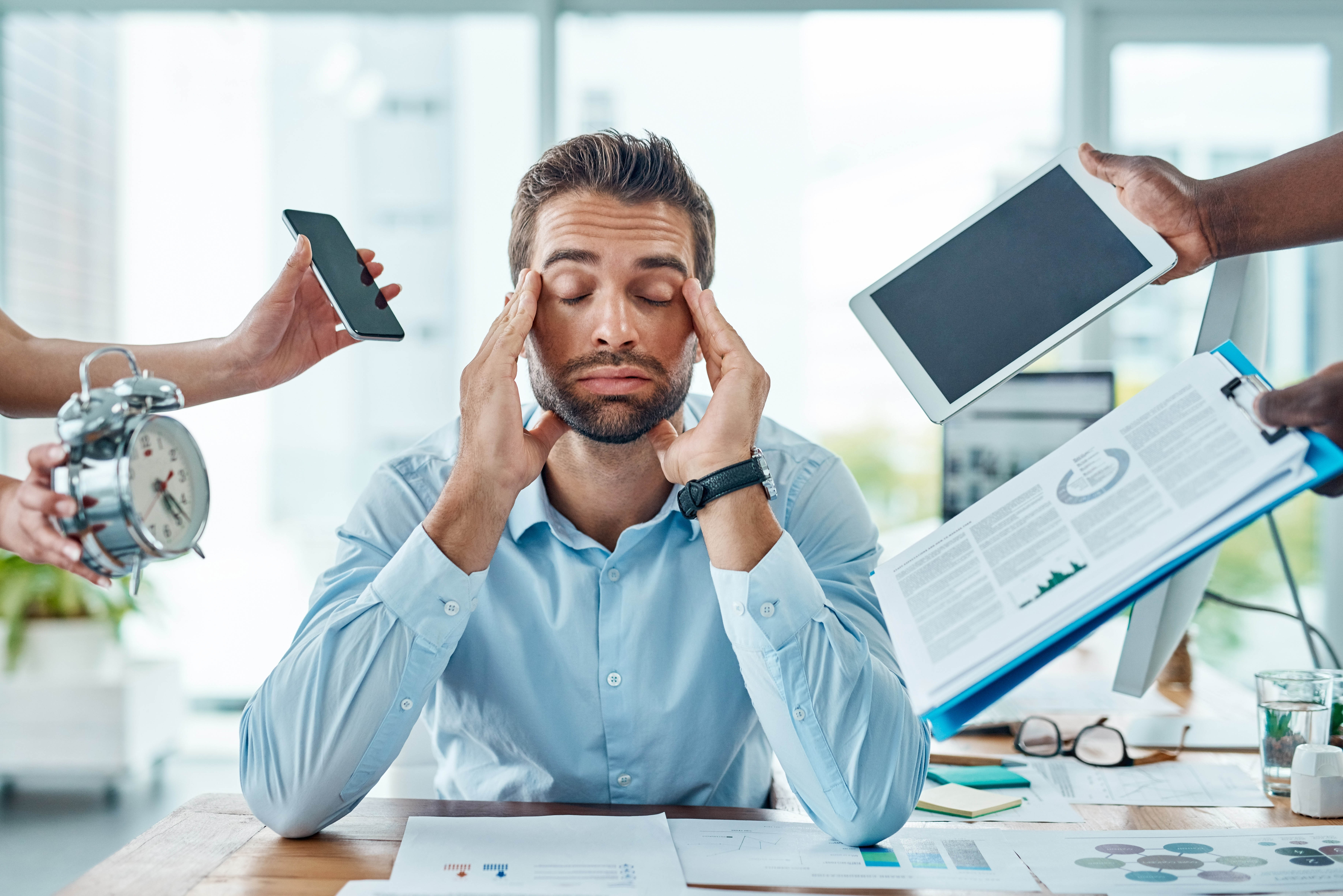 Man suffering from burnout, with other hands thrusting phones, tablets, and more in his face while he has his eyes closed and hands next to his temples